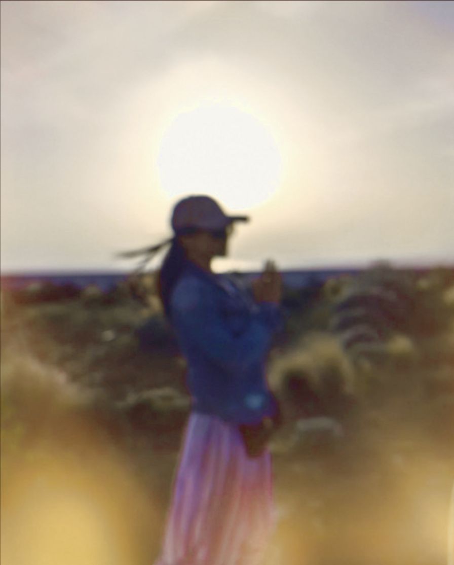 CHELX in a field with the sun behind — a figure in a cap and denim jacket, hair caught by the wind.
